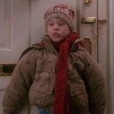 Child wearing a brown coat, red scarf, and patterned hat in front of a door.
